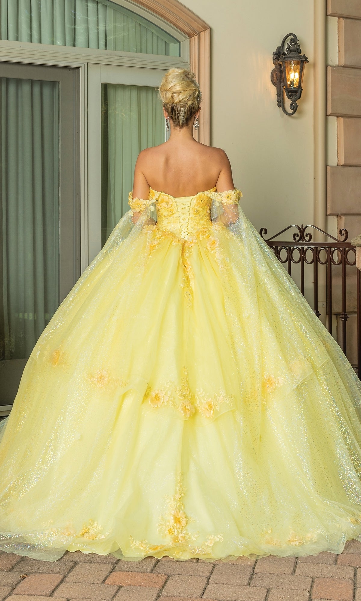 A person in the Quinceanera Dress 1788 By Dancing Queen, featuring a yellow a-line silhouette and off-the-shoulder straps, stands outdoors with their back to the camera near a window and wall-mounted lantern.